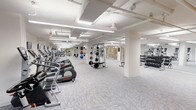 This fitness center offers a comprehensive workout environment designed to accommodate a variety of exercise needs. The space is generously proportioned, with a clean, open layout that allows for easy navigation between different workout zones. Bright, recessed fluorescent lighting illuminates the entire floor, complemented by a crisp white color palette that keeps the atmosphere feeling fresh and energizing.

Along one side of the facility, a long row of cardio equipment — including elliptical trainers and treadmills — lines the wall, providing ample options for aerobic exercise. Toward the center and rear of the space, a dedicated strength training area features a multi-function cable and weight machine, along with a rack of medicine balls for functional fitness. A separate free weights section houses an extensive dumbbell rack, offering a full range of weight options for resistance training.

The flooring throughout consists of durable, patterned fitness carpet tiles that provide both comfort and practicality. Structural columns are integrated seamlessly into the layout without disrupting the flow of the space. Mirrored walls in key sections enhance the sense of openness while allowing users to monitor their form. The overall ambiance is clean, professional, and purpose-built for an effective fitness experience.