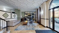 This sophisticated lobby and lounge space showcases a striking combination of classical architecture and modern design. The most commanding feature is the intricate white plasterwork ceiling, adorned with elaborate geometric and floral motifs that add a sense of grandeur throughout the space. Warm travertine tile flooring runs continuously underfoot, complementing the refined aesthetic.

The hallway entry is defined by tall arched windows framed with floor-length charcoal drapery, flooding the space with natural light while offering views to the exterior. Wall sconces provide warm ambient lighting, highlighting the two-tone wall treatment of soft sage green and cream, accented by bold dark molding and decorative diamond-shaped mirror panels.

The living area is thoughtfully arranged with multiple seating vignettes. A central grouping features a wooden coffee table flanked by contemporary grey armchairs atop a blue patterned area rug, while a second cluster near the feature wall pairs cream accent chairs around a round table. A grey sofa and bar-height seating along a built-in counter offer additional options. A glass-partitioned room with dark steel framing provides a semi-private enclosure visible from the main lounge. A staircase with black iron railings connects to upper levels, completing this well-appointed communal space.