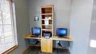 This single-floor business center is designed to meet essential productivity needs in a compact, efficiently arranged space. The focal point is a built-in wooden workstation unit featuring a central tower bookshelf with open shelving for storage and display, flanked by two desk surfaces extending outward on either side. Each desk is equipped with a desktop computer running Windows, a keyboard, and a mouse, providing residents or guests with dedicated workstations. A shared HP laser printer is conveniently positioned at the center of the unit, easily accessible from both seats. Below the central shelving tower, a two-door cabinet offers additional enclosed storage. The walls are painted a neutral gray, contributing to a clean, professional atmosphere. Ergonomic task chairs accompany each workstation, ensuring comfortable use during extended sessions. The flooring features a patterned carpet in muted tones that complements the overall aesthetic. Power outlets are accessible along the wall beneath each desk surface, accommodating various devices. The space is well-lit and organized, making it a practical resource for printing, browsing, or handling everyday business tasks.