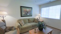 This single-floor living space features a welcoming living room designed with comfort and functionality in mind. The room is finished with light-colored walls and carpeted flooring, contributing to a warm, neutral aesthetic throughout. A plush, sage-green sofa serves as the focal point of the space, accented with decorative patterned throw pillows that add subtle visual interest. A wooden coffee table sits at the center, complemented by a matching side table that holds a classic table lamp, providing soft ambient lighting. A large framed landscape print depicting autumn trees adds a decorative touch to the wall, enhancing the room's inviting character. Natural light enters through a window fitted with horizontal blinds, brightening the space during the day. A potted houseplant positioned near the seating area introduces a natural, organic element to the interior. The overall ambiance is calm and residential, with a layout that makes efficient use of the available space. The single-floor design ensures easy accessibility and a straightforward, cohesive flow throughout the living area.
