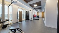 Located at 560 Rideau St in Ottawa, this fitness facility offers a well-appointed exercise space accessed through a connecting hallway. The room is designed with both function and aesthetics in mind, featuring durable rubber flooring throughout that provides traction and comfort during workouts.

The space is divided into distinct workout zones. A strength training area is anchored by a decorative wooden slatted partition wall with dark metal framing, which adds a warm, contemporary architectural element. This zone houses a full dumbbell rack, adjustable weight bench, barbell storage, and a multi-station cable machine. A large wall mirror enhances the sense of openness and supports proper form during training.

The cardio zone faces a bank of large windows that flood the space with natural light and offer elevated views of the Ottawa cityscape — an inspiring backdrop for treadmills, elliptical trainers, a stationary bike, and a recumbent cycle, all from Life Fitness.

The coffered ceiling with dark wood inlays and recessed and pendant lighting adds a refined, upscale character to the room. Subtle cove lighting accents the ceiling perimeter, contributing to a polished ambiance. A stability ball and functional training equipment round out the offerings, making this a comprehensive fitness amenity suited for a wide range of workout routines.