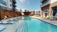 This amenity space at 1425 Semmes Ave in Richmond, Virginia offers a well-appointed fitness center and outdoor pool area designed to serve a contemporary residential community. The fitness center features soaring ceilings that create an open, airy atmosphere, with patterned blue-toned carpet flooring throughout. Floor-to-ceiling glass walls and large steel-framed windows flood the interior with abundant natural light while providing direct views of the outdoor courtyard and surrounding buildings. The space is divided into distinct workout zones separated by glass partition walls with dark metal framing, allowing visual connectivity between areas. Equipment includes cycling bikes, stability balls, strength machines, and open floor space for functional training. A large mounted display screen supports on-demand workout programming. The adjoining outdoor pool area features a generously sized rectangular swimming pool with clear turquoise water, surrounded by a spacious deck furnished with lounge chairs and yellow accent seating. A covered pavilion with a mounted television anchors one end of the pool deck, while the multi-story residential buildings frame the courtyard on multiple sides, creating an enclosed, resort-style setting ideal for relaxation and recreation.