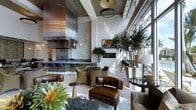 This single-floor space showcases an open-concept design that integrates a sleek kitchen and stylish lounge area within one expansive, flowing layout. The kitchen is anchored by a large island with dark countertops and bar seating, complemented by stainless steel cabinetry, a substantial range hood, and pendant lighting that adds a polished, contemporary feel. Travertine or stone-clad accent walls provide warmth and texture throughout the space, contrasting beautifully with the light hardwood flooring underfoot.

Soaring ceilings amplify the sense of volume, while a dramatic drum pendant fixture serves as a central focal point overhead. A wall of floor-to-ceiling windows lines one side of the space, flooding the interior with natural light and offering views of lush tropical greenery beyond. Tall potted palms and tropical plants bring the outdoors in, enhancing the relaxed yet upscale atmosphere.

The lounge area features rich leather seating and woven accent chairs arranged around low tables, creating an inviting, comfortable gathering space. A wall-mounted flat-screen television and subtle recessed lighting complete the refined, resort-inspired ambiance of this well-appointed space.