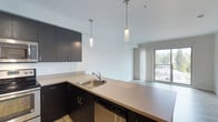 This single-floor space showcases a clean, modern aesthetic with an open-concept layout that seamlessly connects the kitchen and living areas. The kitchen is appointed with dark espresso cabinetry, sleek neutral-toned countertops, a stainless steel sink, and an integrated dishwasher. Two elegant cylindrical pendant lights hang above the kitchen peninsula, providing focused task lighting while adding a stylish design element to the space. The floors feature warm wood-look laminate that runs throughout, complementing the contemporary color palette of deep cabinetry contrasted against bright white walls and ceilings. The living area benefits from abundant natural light streaming through a large floor-to-ceiling window fitted with sheer vertical blinds, which also opens onto what appears to be a balcony with views of the surrounding greenery and streetscape. The high ceilings throughout amplify the sense of openness and airiness. The overall ambiance is bright, clean, and functional — ideal for those who appreciate a contemporary, minimalist living environment with well-considered finishes and a practical, flowing floor plan.