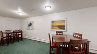 Located at 3415 Federal Dr in Eagan, Minnesota, this shared community space is thoughtfully designed to serve residents with two primary functional areas. The main common room features rich green carpeting, white walls adorned with framed artwork, and baseboard heating that ensures year-round comfort. Multiple dining tables with sturdy wooden chairs provide ample seating, making the space well-suited for casual gatherings or shared dining. Textured ceilings with flush-mount light fixtures keep the atmosphere well-lit and clean throughout. A hallway with a wood-paneled entry door connects the main room to the adjacent areas, maintaining a clear and accessible flow. The dedicated laundry room is equipped with multiple commercial-grade washing machines and dryers, a utility sink, and built-in shelving for added convenience. A small adjoining workspace off the laundry area includes a folding table and seating, ideal for sorting laundry or other tasks. The overall design is practical and straightforward, with neutral tones and durable finishes that prioritize ease of maintenance and everyday usability for residents of the building.