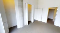 This single-level space includes a carpeted room that serves as a bedroom or flexible living area. A notable architectural detail is the exposed concrete column, which adds an industrial character to the otherwise clean, minimalist interior with white walls. The room features a built-in closet with bi-fold doors, offering shelving and hanging rod storage. Wire shelving is also visible along one wall, providing additional open storage. A second door leads to an adjoining room or hallway, suggesting a connected layout. The neutral-toned carpet and bright white finishes create a simple, functional atmosphere throughout.