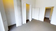 This single-level space includes a carpeted room that serves as a bedroom or flexible living area. A notable architectural detail is the exposed concrete column, which adds an industrial character to the otherwise clean, minimalist interior with white walls. The room features a built-in closet with bi-fold doors, offering shelving and hanging rod storage. Wire shelving is also visible along one wall, providing additional open storage. A second door leads to an adjoining room or hallway, suggesting a connected layout. The neutral-toned carpet and bright white finishes create a simple, functional atmosphere throughout.