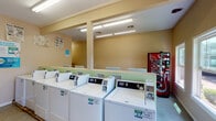 This single-floor shared laundry facility is designed for practical, everyday use and offers a comprehensive set of amenities within a clean, functional layout. The main area is lined with a row of coin-operated top-loading washing machines and dryers, each clearly numbered and equipped with digital controls for ease of use. Instructional signage is posted along the walls, guiding users through washing and drying cycles. Fluorescent lighting runs along the ceiling, ensuring the space is well-lit throughout. A folding table provides a dedicated surface for sorting and folding laundry, while a waste bin nearby keeps the area tidy. A bank of individual mailboxes is integrated into the space, adding a convenient community amenity. A Coca-Cola vending machine is positioned near one wall, offering refreshments during laundry sessions. The walls are finished in a warm beige tone, and the floors feature durable speckled concrete. Natural light filters in through several windows that overlook the surrounding exterior, and a white entry door provides direct outdoor access. A green bulletin board serves as a community information hub. Safety is addressed with a clearly visible fire extinguisher and exit signage. Overall, the facility is straightforward, well-organized, and built to serve the practical needs of a shared residential community.