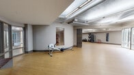 This single-floor fitness studio offers a spacious, open layout designed to accommodate a wide variety of workout styles and training activities. The warm-toned hardwood-style flooring runs throughout, providing both durability and a welcoming aesthetic that complements the neutral gray walls.

The studio is organized into distinct functional zones. One area features a rowing machine and a wall-mounted observation window, creating an engaging and interactive training environment. Suspension training straps are anchored to the wall nearby, supporting bodyweight and functional fitness exercises. A dedicated section houses a rack of weighted medicine balls and stability balls, alongside a punching bag suspended from the ceiling for combat-style training. Stationary cycling bikes occupy another zone, offering cardio training options.

Expansive floor-to-ceiling glass doors along one wall flood the space with natural light and provide views of a landscaped outdoor courtyard, contributing to an open and energizing atmosphere. The exposed ceiling infrastructure, fitted with linear fluorescent lighting, adds an industrial character while ensuring the space is brightly and evenly illuminated throughout.

Audio speakers are mounted near the ceiling, and a wall-mounted control panel supports class or session management. The overall design is clean, functional, and versatile, making this studio well-suited for group fitness classes, personal training, and a broad range of individual workout programs.