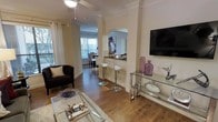 This single-floor residence showcases a thoughtfully designed open-concept layout that promotes a natural flow between the living, dining, and kitchen areas. The living room is anchored by warm hardwood flooring, elegant crown molding, and neutral cream-toned walls that create a bright, inviting atmosphere. A ceiling fan with pendant lighting illuminates the space, while large windows dressed with white curtains and blinds allow generous natural light to filter in.

A defining architectural feature is the arched pass-through opening that connects the kitchen bar area to the living space. The bar counter accommodates stylish high-top stools, making it ideal for casual dining or entertaining. The wall-mounted flat-screen television is complemented by a sleek glass and chrome media console, adding a contemporary touch.

The dining area, visible through the archway, features additional window coverage, maintaining the home's bright and airy character throughout. Decorative accents, including glass vases and curated tabletop accessories, contribute to the polished, cohesive aesthetic. Overall, the space balances classic architectural details with modern furnishings for a refined yet comfortable ambiance.
