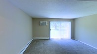 This single-floor residential space offers a clean and functional layout designed for comfortable everyday living. The main living area is finished with wall-to-wall carpeting that adds warmth underfoot, while a textured ceiling provides a classic touch overhead. Neutral-toned walls create a versatile backdrop, making the space easy to personalize.

A full-length sliding glass door serves as a prominent architectural feature, flooding the room with natural light and offering a direct view of lush greenery outdoors. Vertical blinds provide privacy and light control as needed. The expansive glass opening creates a seamless visual connection between the interior and the outdoor environment, enhancing the sense of openness throughout the space.

Climate comfort is well-addressed with both a wall-mounted air conditioning unit and baseboard heating running along the perimeter of the room, ensuring year-round temperature control regardless of the season. The combination of these heating and cooling systems reflects a practical approach to residential comfort.

Overall, the space presents a straightforward, well-lit interior with key functional amenities in place, making it a solid foundation for a comfortable living environment.