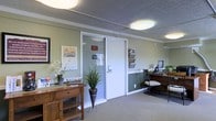 This single-floor professional office space offers a thoughtfully designed interior that balances functionality with a warm, welcoming atmosphere. The reception area serves as the heart of the space, featuring soft sage green walls accented by crisp white crown molding and baseboards that lend a clean, polished look throughout. Carpeted flooring in a neutral tone adds comfort underfoot and contributes to the overall subdued, professional ambiance. A wooden console table near the entrance provides a decorative focal point, accented with fresh floral arrangements and potted greenery that bring a touch of nature indoors. The reception desk area is equipped with a wall-mounted literature organizer, ensuring informational materials are neatly displayed and easily accessible for visitors. A glass-paneled interior door leads to a private office or back workspace, offering visual openness while maintaining a degree of separation. Flush-mount ceiling light fixtures provide consistent, even illumination throughout the space. A dark wood desk and chair in the reception area offer a dedicated workspace for staff. The overall design is practical yet inviting, creating an environment well-suited for client-facing professional services.