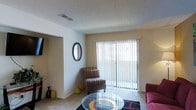 This single-floor residential space offers a functional and comfortable layout designed for everyday living. The living area serves as the heart of the home, featuring soft carpet flooring and a neutral color palette that creates a warm, inviting atmosphere. A large sliding glass door dressed with vertical blinds floods the room with natural light and provides direct access to an outdoor area, offering a seamless connection between interior and exterior spaces. The textured ceiling adds a classic architectural detail throughout. An open passageway connects the living area to an adjacent room, enhancing the sense of flow and accessibility within the floor plan. The living space is well-proportioned, accommodating seating arrangements with ease. Subtle wall decor, including a round mirror, adds a touch of personality to the space. The overall ambiance is bright and airy, with ample natural light filtering through the large sliding door. This single-story layout provides convenient, step-free living throughout, making it practical and easy to navigate from room to room.