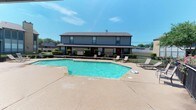 This residential community centers around a spacious, inviting outdoor pool area that serves as the heart of the property. The pool itself is generously proportioned with a geometric shape, featuring clearly marked depth indicators and a clean concrete surround equipped with lounge chairs for relaxing under the open sky. The crystal-clear turquoise water is well-maintained, reflecting the bright sunlight that bathes the area throughout the day.

Surrounding the pool, a wrought iron perimeter fence encloses the space, providing both security and a defined boundary for the amenity area. Neatly trimmed hedges and mature trees add greenery and shade, enhancing the overall ambiance of the outdoor environment.

Adjacent to the pool, a brick-constructed covered pavilion with a hip roof offers a shaded retreat, complete with seating and a vending machine — ideal for gatherings and leisure. The patio extends outward, providing ample space for outdoor furniture and social activities.

The multi-story residential buildings visible in the background feature gray vinyl siding, brick accents, dark shingled rooflines, and red entry doors, giving the community a cohesive and classic architectural character. The grounds are well-kept, with maintained landscaping throughout, contributing to a comfortable and community-oriented living environment.