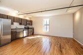 This single-floor loft-style unit offers a well-designed layout that balances industrial charm with modern functionality. Warm hardwood floors run throughout the main living areas, complementing the exposed wood structural beams and columns that serve as standout architectural features. Track lighting runs along the ceiling, providing flexible, well-distributed illumination across the open living space.

The kitchen is efficiently appointed with rich dark wood cabinetry, light countertops, and a full suite of stainless steel appliances including a side-by-side refrigerator, electric range, built-in microwave, and dishwasher. The layout maximizes storage and counter space within the compact footprint.

A hallway lined with warm wood-toned doors connects the main living area to the bathroom and additional rooms. The bathroom features neutral stone-look tile flooring, a wood-toned vanity with ample cabinet storage, a white countertop, a large mirror with overhead lighting, and a full toilet and towel bar.

Exposed ductwork along the ceiling adds to the loft's industrial aesthetic, while crisp white walls keep the space feeling bright and open. The overall ambiance is one of urban character paired with practical, contemporary finishes — making this a distinctive and functional living space.