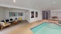 Located at 12224 Daphne Ln NW in Silverdale, Washington, this single-level indoor pool facility offers a well-appointed aquatic and relaxation space designed for year-round use. The centerpiece is a generously sized indoor swimming pool with lane markers, complemented by an adjoining octagonal spa/hot tub, both finished with light blue tile work that enhances the bright, clean aesthetic of the space.

The interior is characterized by crisp white walls, recessed lighting, and smooth concrete-style flooring that extends throughout. Large windows and multiple glass-panel doors flood the space with natural light while framing views of the surrounding mature landscaping and outdoor patio areas. Several door access points connect the interior seamlessly to exterior lounge spaces, creating a fluid indoor-outdoor experience.

A dedicated lounge area features a circular upholstered daybed with an array of decorative pillows, accent chairs, and casual seating, providing a comfortable retreat alongside the pool. Colorful artwork adorns the walls in a secondary seating nook near what appears to be a sauna or changing room access. Safety equipment, including a life ring and fire extinguisher, is visibly and conveniently placed throughout the space. The overall ambiance is modern, airy, and resort-inspired.