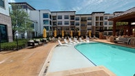 Located at 351 Northwood Dr in Flower Mound, Texas, this single-level outdoor amenity space is designed around a generously sized swimming pool that serves as the centerpiece of the community's exterior. The pool features a curved, freeform design complemented by a shallow entry area, with stainless steel entry rails providing easy access. The surrounding deck is finished in warm-toned stamped concrete, offering ample space for lounging and circulation.

A standout feature of the space is the covered brick pavilion situated at one end of the pool. Built with a pitched metal roof and supported by brick columns, it houses an outdoor seating and dining area, complete with a mounted television, making it well-suited for social gatherings. Additional seating is scattered throughout the deck, including white chaise lounges and accent chairs paired with yellow market umbrellas that add a vibrant contrast to the neutral palette.

Adjacent covered patios with glass sliding doors and modern outdoor furniture provide semi-private retreat options overlooking the pool. The surrounding multi-story brick and stucco buildings frame the courtyard, contributing to an enclosed, resort-like ambiance. Tall cypress trees and landscaped greenery add vertical interest and a sense of privacy throughout the space.
