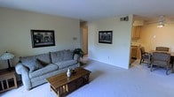 This single-floor space presents a clean, functional layout centered around a well-proportioned living area. The room is finished with wall-to-wall carpet in a neutral tone, contributing to a warm and understated ambiance throughout. Soft, cream-colored walls provide a bright backdrop, keeping the space feeling open and airy.

The living area is arranged around a gray upholstered sofa accented with decorative pillows, paired with a solid wood Mission-style coffee table featuring drawer storage — a practical yet classic centerpiece. Two framed black-and-white artworks are displayed on the walls, adding visual interest without overwhelming the space. A potted plant in the corner introduces a touch of greenery.

A wall-mounted HVAC unit provides climate control, and the ceiling is clean and unobstructed. A doorway visible along the back wall leads to additional areas of the unit. To the right, the living space opens directly into the kitchen, where light wood cabinetry and appliances are partially visible, suggesting a convenient and connected layout. The overall atmosphere is modest, tidy, and move-in ready.