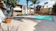 This outdoor amenity space showcases a well-appointed pool area designed in a warm, Southern California aesthetic. The centerpiece is a generously sized swimming pool with clear turquoise water, complemented by a separate circular spa/hot tub nearby. The surrounding deck is finished in light-toned concrete, providing ample space for lounging, and is lined with rows of sling-style chaise lounges ideal for sunbathing.

The architecture bordering the pool area reflects a classic Spanish-influenced style, characterized by stucco exteriors in warm tan and cream tones, clay tile rooflines, and wood lattice privacy screens on the balconies. Tall palm trees and mature shade trees are interspersed throughout the grounds, adding a tropical ambiance and natural privacy screening.

A covered patio area adjacent to the pool deck features exposed wood beam ceilings, outdoor table-and-chair seating, and vending machines for added convenience. This shaded retreat offers a comfortable space to relax out of direct sunlight. A wrought iron security gate provides controlled access to the pool area, enhancing privacy and safety. The overall atmosphere is relaxed and resort-like, suited for outdoor recreation and leisure in a sunny climate.