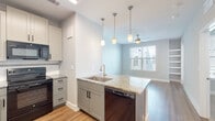 This well-appointed single-floor unit offers a clean, modern aesthetic defined by a neutral color palette, wood-look flooring, and an abundance of natural light throughout. The open-concept kitchen is a standout feature, centered around a peninsula island with granite countertops, an undermount sink with a brushed gold faucet, and elegant pendant lighting overhead. The kitchen is fully equipped with black stainless appliances, including a range, built-in microwave, and dishwasher, all complemented by crisp white shaker cabinetry and a subway tile backsplash. The kitchen flows seamlessly into the living and dining area, which benefits from natural light through blinds-fitted windows and is finished with a ceiling fan for added comfort. Built-in shelving along one wall provides practical storage and display space. The bedroom features plush carpet flooring, a ceiling fan, and a window with exterior views, creating a comfortable and restful retreat. A second carpeted room offers additional flexible living space. The bathroom is finished with large-format light tile in the tub/shower surround, wood-look flooring, and matching granite countertops, maintaining the cohesive design language found throughout the unit. Overall, this space presents a polished, move-in-ready environment with thoughtful finishes and a functional layout.