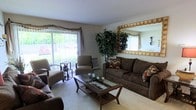 This single-floor residence offers a comfortable and thoughtfully arranged living space designed for both relaxation and everyday functionality. The living room serves as the centerpiece of the home, featuring a generously sized sofa and coordinating armchairs upholstered in rich, dark fabric, complemented by decorative accent pillows. A glass-topped coffee table with a wooden frame anchors the seating arrangement, adding an elegant touch to the room.

Natural light flows abundantly through large sliding glass doors dressed with vertical blinds, offering a view of the lush greenery outside and creating a seamless connection between the interior and the outdoors. The soft, light-toned carpeting underfoot enhances the room's warm and cozy atmosphere.

A tall, full indoor tree plant adds a refreshing natural element to the space, bringing life and color to the interior. A generously sized decorative mirror with a gilded frame is mounted on the wall, visually expanding the room and adding a classic, refined detail. The neutral wall tones throughout provide a versatile backdrop that ties the room's furnishings and accents together cohesively. Overall, this residence presents a well-balanced blend of comfort, natural light, and tasteful design.