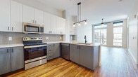 This contemporary single-floor residence showcases a thoughtfully designed open-concept layout with warm hardwood flooring flowing throughout the main living areas. The kitchen is a standout feature, offering two-tone cabinetry with white upper cabinets and gray lower cabinets, stainless steel appliances including a range, microwave, dishwasher, and refrigerator, and a peninsula with a matte black sink and pendant lighting overhead. A built-in wine rack and open shelving add both function and character to the adjacent dining or bar area, complete with tile backsplash accents. Large floor-to-ceiling windows allow abundant natural light and provide views of the surrounding cityscape, creating a bright and airy atmosphere throughout the living space. The bedroom is finished with plush carpeting and features a generously sized window framing expansive city views, filling the room with daylight. The bathroom is clean and modern, with large-format white subway tile surrounding a soaking tub and shower combo, black hardware accents, and light wood-look tile flooring. A dedicated laundry area houses a stacked full-size washer and dryer unit, adding everyday convenience. The overall design balances modern industrial touches with warm, residential finishes.