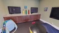 Kids Kave is a cheerfully designed children's space crafted to provide a fun, engaging environment for young visitors. The room is finished with soft sage-green walls that create a calm yet playful atmosphere, complemented by durable dark flooring well-suited for active play.

A standout feature of the space is the long built-in cabinetry running along one wall, finished in warm wood tones with a granite-style countertop. Two flat-screen televisions are mounted above the cabinetry, offering ample entertainment options. Oversized decorative pencil figures in bold primary colors — yellow, green, and red — are displayed on the counter, while matching pencil wall décor adds to the creative, school-inspired theme. Colorful educational artwork, including number charts and vibrant paintings, is displayed throughout the room to stimulate young minds.

A chalkboard mounted on one wall invites hands-on creativity, while a glass-paneled door connects the space to an adjacent area, providing natural sightlines for supervision. A bin of plush toys and a children's play tent add interactive play elements to the room. The overall design of Kids Kave balances functionality and fun, making it an ideal dedicated space for children.
