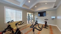 This single-floor spin studio is purpose-built for indoor cycling workouts and offers a well-appointed, focused fitness environment. The room accommodates multiple Matrix-brand stationary spin bikes arranged to provide ample space for each user, making it suitable for both individual sessions and group cycling classes. A large wall-mounted flat-screen television is positioned at the front of the room, paired with a soundbar, offering an engaging visual focal point during workouts. An additional smaller display is mounted nearby, likely for scheduling or instructional use. The architectural design features vaulted tray ceilings with recessed lighting, lending the studio a sense of height and openness. The soft gray walls are complemented by white crown molding and baseboards, contributing to a clean, modern aesthetic. Patterned carpeting with warm orange accent tones covers the floor, adding a dynamic visual element to the space. Natural light enters through multiple windows fitted with horizontal blinds, keeping the environment bright and welcoming. Glass sliding doors connect the spin studio directly to a larger main fitness area, visible in the background and equipped with treadmills and strength training machines, allowing seamless access to a full range of fitness amenities.