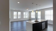 This single-floor space showcases a thoughtfully designed open-concept layout that seamlessly connects the kitchen and living areas. The kitchen serves as a central focal point, anchored by a sleek island with a white countertop, an integrated sink, and dark cabinetry that creates a striking contrast against the lighter surroundings. Three stylish pendant lights hang above the island, providing warm, focused illumination that enhances the modern aesthetic. Recessed lighting throughout the ceiling further brightens the space, complementing the ample natural light that pours in through multiple windows dressed with horizontal blinds. The living area flows effortlessly from the kitchen, offering an open and airy feel underscored by smooth hardwood-style flooring in a cool gray tone that extends throughout. Neutral gray walls and crisp white trim lend a clean, contemporary look, while the open floor plan creates a sense of spaciousness and flexibility. The abundant windows on multiple walls ensure the interior remains bright and connected to the outdoors. Overall, the space combines modern design elements with a welcoming, functional layout suited for everyday living.