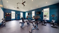 Located at 8352 E Broad St in Blacklick, Ohio, this single-floor fitness center is a well-appointed facility designed to support a comprehensive workout experience. The interior is defined by bold teal-blue accent walls contrasted against crisp white trim and ceilings, creating an energizing and modern atmosphere throughout.

The main workout area is generously sized and features durable rubber flooring, recessed lighting, and ceiling fans that promote air circulation. Multiple large windows line the walls, flooding the space with abundant natural light and offering views of the surrounding greenery. Two wall-mounted flat-screen televisions are positioned for easy viewing from cardio equipment.

The cardio section is equipped with a row of treadmills, stationary bikes including recumbent and upright models, an elliptical trainer, and a stair climber, providing a full range of cardiovascular training options. A dedicated strength training area houses a power rack with a barbell, a multi-functional cable machine, dumbbells, kettlebells, medicine balls, and TRX suspension straps — catering to both free weight and functional fitness routines.

A separate transitional area features vaulted ceilings, a stainless steel water fountain, and an additional cable/functional trainer machine, providing a practical and convenient space between the main workout zones.