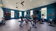 Located at 8352 E Broad St in Blacklick, Ohio, this single-floor fitness center is a well-appointed facility designed to support a comprehensive workout experience. The interior is defined by bold teal-blue accent walls contrasted against crisp white trim and ceilings, creating an energizing and modern atmosphere throughout.

The main workout area is generously sized and features durable rubber flooring, recessed lighting, and ceiling fans that promote air circulation. Multiple large windows line the walls, flooding the space with abundant natural light and offering views of the surrounding greenery. Two wall-mounted flat-screen televisions are positioned for easy viewing from cardio equipment.

The cardio section is equipped with a row of treadmills, stationary bikes including recumbent and upright models, an elliptical trainer, and a stair climber, providing a full range of cardiovascular training options. A dedicated strength training area houses a power rack with a barbell, a multi-functional cable machine, dumbbells, kettlebells, medicine balls, and TRX suspension straps — catering to both free weight and functional fitness routines.

A separate transitional area features vaulted ceilings, a stainless steel water fountain, and an additional cable/functional trainer machine, providing a practical and convenient space between the main workout zones.