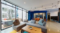 This single-floor communal space makes a bold first impression with a colorful, large-scale mural spanning the entrance lobby wall, set against polished concrete floors and a cobalt blue ceiling with sleek linear lighting. A dedicated package and mailbox area adds practical functionality near the entry.

The main lounge area is anchored by floor-to-ceiling black-framed windows that flood the space with natural light and provide direct views of an outdoor courtyard patio. Modular gray sectional sofas, a warm wood coffee table, and a vibrant multicolored area rug create an inviting, contemporary gathering area. A wall-mounted flat-screen television and open industrial-style shelving complete the lounge.

Adjacent to the lounge, a dining area features a long communal table paired with bold orange chairs, set against a deep cobalt blue accent wall. A glass-partitioned room beyond provides an additional enclosed gathering or workspace.

The fully equipped kitchen is sleek and modern, showcasing high-gloss white cabinetry, stainless steel appliances, a generous island with a white quartz countertop, undermount sink, and pendant lighting. Throughout, polished concrete floors and vibrant color accents unify the space with a cohesive, energetic aesthetic.