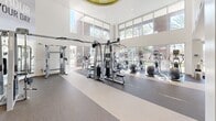 Located at 5805 Charlotte Dr in San Jose, CA, this modern fitness center offers a spacious, well-appointed workout environment spread across a single floor. The facility is divided into two primary zones, each designed to support a comprehensive fitness experience.

The main cardio and strength area is defined by soaring double-height ceilings and an expansive wall of floor-to-ceiling windows that flood the space with natural light and provide open views of the surrounding palm tree-lined exterior. Rows of elliptical trainers, treadmills, and stationary bikes line the perimeter, while a multi-station cable and weight machine system anchors the center of the floor. Bold motivational wall graphics and branding add an energetic visual character to the space.

A second, more intimate workout zone features dedicated strength training equipment, a full dumbbell rack, Precor selectorized machines, and mirrored walls that visually expand the room. Circular recessed ceiling fixtures and ceiling fans provide comfortable, well-distributed lighting throughout. Durable rubber flooring throughout both areas ensures safety and functionality.

Together, the two zones create a cohesive, professional-grade fitness facility that balances high energy with practical design, making it well-suited for a wide range of workout routines and fitness levels.