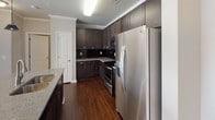 This thoughtfully designed single-floor residence offers a seamless blend of comfort and modern style across its well-proportioned living spaces. The open-concept kitchen serves as a central highlight, boasting rich dark cabinetry, granite countertops, a large breakfast bar with seating, stainless steel appliances including a range, microwave, and side-by-side refrigerator, and warm hardwood-style flooring. Pendant lighting adds a contemporary touch above the kitchen bar, while crown molding throughout lends an air of refinement. A dedicated laundry closet with stacked washer and dryer units is conveniently accessed directly from the kitchen area. The living room is inviting and comfortable, featuring plush carpeting, crown molding, a ceiling fan with light, and access to an outdoor area through glass-paned doors that bring in generous natural light. The bedroom is a calm, restful retreat with soft carpet, a ceiling fan, and neutral tones that create a relaxed ambiance. The bathroom is cleanly finished with tile flooring, a dark-cabineted vanity with granite countertop, and a large mirror. Throughout the space, a cohesive neutral color palette ties each room together, creating a harmonious and welcoming environment.