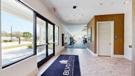 Located at 2600 Lake Betty Blvd in Apopka, Florida, this single-story amenity facility at BLVD 2600 Luxurious Apartment Living spans a generous footprint across two distinct areas — a welcoming lounge and a fully equipped fitness center. The entrance greets visitors with large black-framed glass doors, stone-look tile flooring, and a bold branded welcome mat, setting an upscale tone from the start. A geometric diamond pendant light fixture accents the entryway corridor, leading seamlessly into the facility's two zones. The lounge area features warm lighting, built-in cushioned bench seating upholstered in golden tones, and curated wall art, including a large world map print, creating a comfortable and stylish gathering space. The fitness center is the standout feature of the space, boasting soaring vaulted ceilings with exposed wood beam accents and a large ceiling fan for air circulation. A vivid geometric-patterned carpet in blue and grey tones covers the floor, adding a modern design element throughout. Walls of windows on multiple sides flood the space with natural light and offer views of the surrounding outdoor landscape. Equipment includes a Synergy 360 functional training tower, cable machines, strength benches, treadmills, rowing machines, free weights, and cardio equipment. Full-length mirrors and warm wood-paneled wall accents complete the polished, high-end aesthetic.