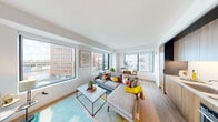 This contemporary single-floor residence is designed with a clean, modern aesthetic throughout. The open-plan living and dining area is the heart of the space, characterized by large floor-to-ceiling windows on multiple walls that flood the interior with natural light and frame sweeping views of the surrounding urban skyline. Light hardwood flooring runs continuously through the main living areas, complementing the bright white walls and ceilings.

The kitchen is efficiently designed with full-height wood-grain cabinetry in a warm oak finish, integrated black hardware, a white countertop, built-in oven, cooktop, and a recessed microwave — offering a sleek, organized cooking environment. The dining nook sits adjacent, positioned beside a wide corner window that offers an elevated city panorama.

The bedroom is a calm retreat featuring a window with city views, ample wall space, and direct access to the living areas. Two bathrooms serve the residence: one features a dark stone-topped vanity with a wood-grain base, an illuminated mirror cabinet, and modern fixtures; the second is a full bath with a soaking tub, a coordinating vanity, and tile flooring. Together, the rooms present a cohesive, thoughtfully designed urban living space with strong architectural detail and quality finishes throughout.