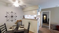 This single-story space showcases a practical and well-connected open layout that promotes a seamless flow between living areas. The dining area is positioned adjacent to the kitchen, separated by a half-wall pass-through that features a built-in ledge — ideal for casual serving or additional counter space. This design element keeps the areas visually connected while maintaining a defined separation between the two spaces.

The kitchen is equipped with white cabinetry, white appliances including a range and microwave, and a decorative tile backsplash that adds a subtle textural detail. The space is efficiently arranged for everyday functionality.

The dining area accommodates a dark-toned table and chair set and is complemented by a ceiling fan with a light fixture overhead. Decorative wall art adds a personalized touch to the neutral, light-toned walls. A textured ceiling runs throughout the space.

Hardwood-style flooring extends through the main areas, transitioning to carpeting in the living room visible in the background. The hallway leading further into the unit suggests additional rooms beyond the primary living areas, contributing to the overall sense of a functional, well-organized floor plan.