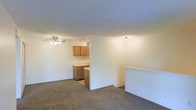 This single-floor space features an open-concept layout that seamlessly connects the living area and kitchen. The living room is finished with wall-to-wall carpeting and a ceiling fan with integrated lighting, providing both comfort and air circulation. A textured ceiling adds a subtle design detail throughout the space.

The kitchen is positioned adjacent to the living area and is partially separated by a half-wall that doubles as a breakfast bar or pass-through counter, enhancing the open feel while defining each functional zone. The kitchen is equipped with warm-toned wooden cabinetry offering ample storage, light-colored countertops, and a sink positioned beneath the upper cabinets. A flush-mount light fixture illuminates the kitchen area.

The neutral color palette of white and beige walls, combined with the light carpeting, creates a clean and bright atmosphere throughout. The layout is practical and efficient, making good use of the available space. The open connection between the kitchen and living area promotes a sociable, accessible flow suited to everyday living.