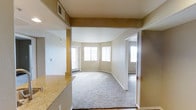 This single-floor residence offers a thoughtfully arranged open-concept layout that connects the kitchen and living areas seamlessly. The kitchen features granite countertops with a warm, neutral tone, providing both functionality and a polished aesthetic. A raised counter bar separates the kitchen from the main living space, ideal for casual dining or entertaining.

The living area is carpeted in a neutral tone, contributing to a comfortable and inviting atmosphere. Its curved wall design adds an architectural touch that softens the overall feel of the space. Multiple windows allow abundant natural light to flow through, brightening the interior throughout the day. Vertical blinds on the windows offer flexible light and privacy control.

Warm, earth-toned walls in shades of tan and beige run throughout the unit, creating a cohesive and calming color palette. Visible HVAC vents in the ceiling indicate a central air system for year-round climate comfort. A doorway visible from the main living area leads to an additional room, suggesting a separate bedroom or den with hardwood-style flooring. White baseboards and trim provide clean contrast against the neutral walls, completing the well-maintained interior of this functional and comfortable single-level home.