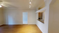 This single-floor space showcases a thoughtfully designed open layout that seamlessly connects the living area and kitchen. The living area is finished with warm hardwood flooring that extends throughout, complemented by crisp white baseboards and elegant crown molding that frame the room with a clean, polished look. A track lighting system mounted on the ceiling provides adjustable, directed illumination across the space.

A defining architectural feature is the kitchen pass-through, which opens the kitchen to the living area, promoting an open and connected feel. The pass-through is topped with dark granite countertops and includes a stainless steel sink, while warm wood cabinetry is visible beyond. Decorative column detailing accents the transition between the kitchen and living space, adding a subtle architectural touch.

The neutral wall tones throughout create a bright, versatile backdrop that enhances the sense of openness. A doorway visible from the main living area leads to an adjoining room, suggesting additional functional space beyond. The overall ambiance is clean, modern, and well-appointed, with quality finishes that elevate the everyday living experience.