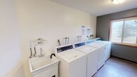 This single-floor laundry facility is designed for practical, efficient use. The room is laid out in a straightforward, functional arrangement, with a row of top-loading washing machines positioned along the main wall. Each machine is equipped with its own dedicated water hookup and shut-off valves, ensuring organized and independent operation. The machines feature digital control panels, indicating modern, user-friendly functionality.

To the left of the washing machines sits a white utility sink mounted on a pedestal, complete with a standard faucet, providing a convenient hand-washing or pre-treatment station. Informational notices are posted on the wall above the machines, offering usage guidelines for occupants.

The room's interior is finished with clean white walls and light-toned flooring, contributing to a bright and hygienic atmosphere. The neutral color palette and uncluttered layout keep the space feeling orderly and easy to navigate. The overall design prioritizes utility and cleanliness, making it a well-equipped shared laundry space suited to accommodate multiple users efficiently.