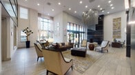 This elegantly designed single-floor space presents a seamless blend of contemporary and transitional styling, anchored by dramatically soaring ceilings that create an impressive sense of volume and grandeur. The centerpiece of the main lounge area is a striking cylindrical chandelier composed of cascading metallic chains, drawing the eye upward and commanding attention from every vantage point.

The open-plan layout flows effortlessly between distinct functional zones. A generously sized lounge area is furnished with a rich leather Chesterfield sofa, tufted ottoman, and an arrangement of neutral upholstered armchairs atop a patterned area rug with gold accents. A formal dining area nearby features a large dark-wood table with seating for eight, topped by an ornate crystal chandelier, set against floor-to-ceiling windows that invite abundant natural light and views of the surrounding streetscape.

A service counter with dark cabinetry, a wall-mounted display screen, and under-counter appliances provides practical amenities. Adjacent corridors are tastefully appointed with curated wall art and tile flooring. Dark accent walls with horizontal paneling, mirrored upper-level railings, and recessed lighting throughout contribute to the space's polished, upscale ambiance.