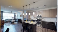 This single-floor residence in Little Elm offers a thoughtfully designed open-concept layout that seamlessly connects the kitchen, dining, and living areas into one cohesive living space. The kitchen serves as the centerpiece, featuring sleek two-toned cabinetry in light and dark finishes, stainless steel appliances including a range, microwave, and dishwasher, and a granite-topped island with bar seating for casual dining. A trio of pendant lights hang above the island, complementing the track lighting overhead and creating a warm, layered ambiance throughout. Rich dark hardwood flooring runs throughout the space, adding depth and contrast to the light cabinetry and neutral walls. The open living area beyond the kitchen benefits from natural light filtering through large windows dressed with soft drapery, offering a comfortable and inviting atmosphere. The fluid connection between the kitchen, dining space, and living room makes this residence well-suited for both everyday living and entertaining. The overall design aesthetic is modern and clean, with contemporary finishes and fixtures that contribute to a polished, cohesive look throughout the home.