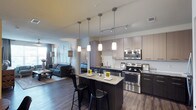 This single-floor residence in Little Elm offers a thoughtfully designed open-concept layout that seamlessly connects the kitchen, dining, and living areas into one cohesive living space. The kitchen serves as the centerpiece, featuring sleek two-toned cabinetry in light and dark finishes, stainless steel appliances including a range, microwave, and dishwasher, and a granite-topped island with bar seating for casual dining. A trio of pendant lights hang above the island, complementing the track lighting overhead and creating a warm, layered ambiance throughout. Rich dark hardwood flooring runs throughout the space, adding depth and contrast to the light cabinetry and neutral walls. The open living area beyond the kitchen benefits from natural light filtering through large windows dressed with soft drapery, offering a comfortable and inviting atmosphere. The fluid connection between the kitchen, dining space, and living room makes this residence well-suited for both everyday living and entertaining. The overall design aesthetic is modern and clean, with contemporary finishes and fixtures that contribute to a polished, cohesive look throughout the home.