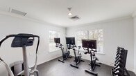 Located at 2323 Downing Ave in Savannah, Georgia, this single-floor fitness facility is thoughtfully designed to support a well-rounded workout experience. The space is divided into distinct exercise zones, each finished with neutral gray flooring, crisp white walls, and crown molding details that lend a clean, polished aesthetic throughout.

The cycling studio features a row of stationary bikes equipped with individual display screens, allowing users to engage in interactive or guided cycling sessions. Multiple windows fitted with white horizontal blinds line the walls, filling the room with natural light and offering views of the surrounding greenery outside.

An adjacent strength and cardio area houses a multi-station cable and weight machine alongside a recumbent bike, an elliptical-style machine, and additional resistance equipment. A wall-mounted fan provides ventilation, and a window allows natural light to complement the overhead lighting in this zone.

A separate section of the facility is dedicated to free weight training, featuring a dumbbell rack stocked with a full range of weights. This area also benefits from ample natural light through multiple windows, maintaining the bright and airy atmosphere consistent across the entire facility.

A glass-panel interior door connects the fitness space to an adjoining area, enhancing the open, accessible flow of the layout. Overall, the facility offers a comprehensive, well-equipped environment suited for a variety of fitness routines.