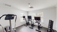 Located at 2323 Downing Ave in Savannah, Georgia, this single-floor fitness facility is thoughtfully designed to support a well-rounded workout experience. The space is divided into distinct exercise zones, each finished with neutral gray flooring, crisp white walls, and crown molding details that lend a clean, polished aesthetic throughout.

The cycling studio features a row of stationary bikes equipped with individual display screens, allowing users to engage in interactive or guided cycling sessions. Multiple windows fitted with white horizontal blinds line the walls, filling the room with natural light and offering views of the surrounding greenery outside.

An adjacent strength and cardio area houses a multi-station cable and weight machine alongside a recumbent bike, an elliptical-style machine, and additional resistance equipment. A wall-mounted fan provides ventilation, and a window allows natural light to complement the overhead lighting in this zone.

A separate section of the facility is dedicated to free weight training, featuring a dumbbell rack stocked with a full range of weights. This area also benefits from ample natural light through multiple windows, maintaining the bright and airy atmosphere consistent across the entire facility.

A glass-panel interior door connects the fitness space to an adjoining area, enhancing the open, accessible flow of the layout. Overall, the facility offers a comprehensive, well-equipped environment suited for a variety of fitness routines.