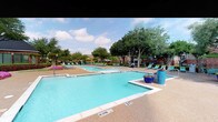 This outdoor amenity space in Frisco, Texas offers an impressive resort-style pool complex designed for relaxation and recreation. The area features multiple swimming pools, including a generously sized lap pool and additional leisure pools, all filled with crystal-clear blue water. A dedicated hot tub/spa provides a separate space for hydrotherapy and unwinding. The pool deck is finished with an aggregate surface, providing both durability and visual appeal, and is lined with rows of vibrant teal and green lounge chairs, inviting sunbathers to spread out comfortably. Colorful potted flowers in bold blue and white planters add decorative accents throughout. Shade umbrellas in bright green are positioned strategically around the deck, offering relief from the Texas sun. The entire complex is enveloped by mature trees and well-maintained landscaping, including blooming pink and purple flowers that add a lively splash of color. In the background, a classic brick clubhouse with a steeply pitched dark roof and large windows provides an attractive architectural backdrop. Outdoor grilling stations are also visible near the clubhouse, enhancing the space's entertainment value. The open-sky setting allows for abundant natural light throughout the day.