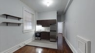 This single-floor space showcases a thoughtfully arranged open layout, with the kitchen serving as a central feature. The kitchenette is equipped with stainless steel appliances, including a full-size refrigerator and a gas range, complemented by dark espresso-toned cabinetry above and below a clean white countertop with an integrated sink. A built-in range hood is installed beneath the upper cabinets, adding functional utility to the cooking area.

The flooring transitions between rich, dark hardwood and light tile in the kitchen zone, creating a natural visual boundary within the open space. Soft grey walls lend a neutral, modern aesthetic throughout. A window fitted with white blinds allows natural light to filter into the kitchen area, brightening the space during daylight hours. White baseboards provide a clean contrast against the darker flooring.

A hallway visible in the background leads to additional rooms, suggesting further private living areas beyond the main open space. The overall ambiance is clean, minimal, and functional, making efficient use of the available footprint.