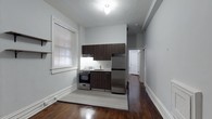 This single-floor space showcases a thoughtfully arranged open layout, with the kitchen serving as a central feature. The kitchenette is equipped with stainless steel appliances, including a full-size refrigerator and a gas range, complemented by dark espresso-toned cabinetry above and below a clean white countertop with an integrated sink. A built-in range hood is installed beneath the upper cabinets, adding functional utility to the cooking area.

The flooring transitions between rich, dark hardwood and light tile in the kitchen zone, creating a natural visual boundary within the open space. Soft grey walls lend a neutral, modern aesthetic throughout. A window fitted with white blinds allows natural light to filter into the kitchen area, brightening the space during daylight hours. White baseboards provide a clean contrast against the darker flooring.

A hallway visible in the background leads to additional rooms, suggesting further private living areas beyond the main open space. The overall ambiance is clean, minimal, and functional, making efficient use of the available footprint.