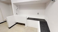 This purpose-built dog washing station is a clean, efficiently designed facility tailored for pet care. The space is finished with bright white walls and white beadboard paneling, giving it a crisp, sanitary aesthetic. At its center is a raised, enclosed wash basin fitted with a chrome handheld showerhead and adjustable water controls, making bath time accessible and convenient for pets of varying sizes. A dark speckled granite-style surface wraps around the basin, providing a durable and non-slip area for pets to safely step and rest. The concrete flooring is easy to clean and built to withstand heavy use. Overhead, exposed silver ventilation ductwork runs prominently across the ceiling, ensuring proper airflow and moisture management within the space — an essential feature in a wet-use environment. A utility area near the entry door houses visible plumbing infrastructure and water supply components, reflecting the functional, no-frills mechanical design of the facility. A prominently displayed Dog Wash Station Guidelines sign is mounted near the entrance, providing users with clear rules for safe and considerate use of the space. Overall, the station is a well-organized, practical amenity designed to make pet washing hygienic and straightforward.